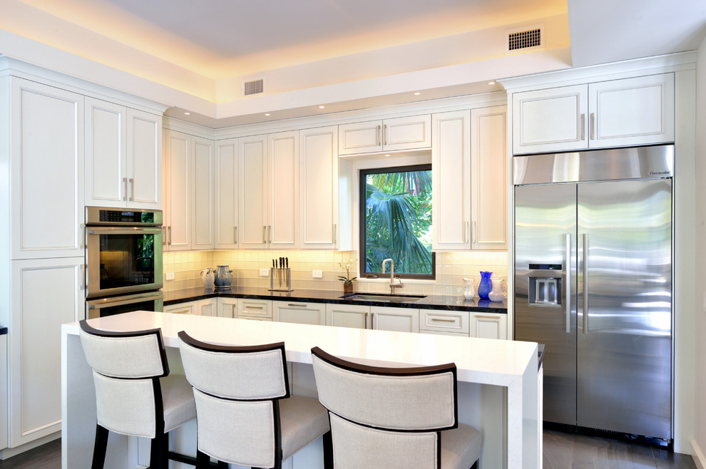 Photo of a large contemporary l-shaped kitchen/diner in Miami with an island, a submerged sink, recessed-panel cabinets, white cabinets, engineered stone countertops, white splashback, glass tiled splashback and stainless steel appliances.