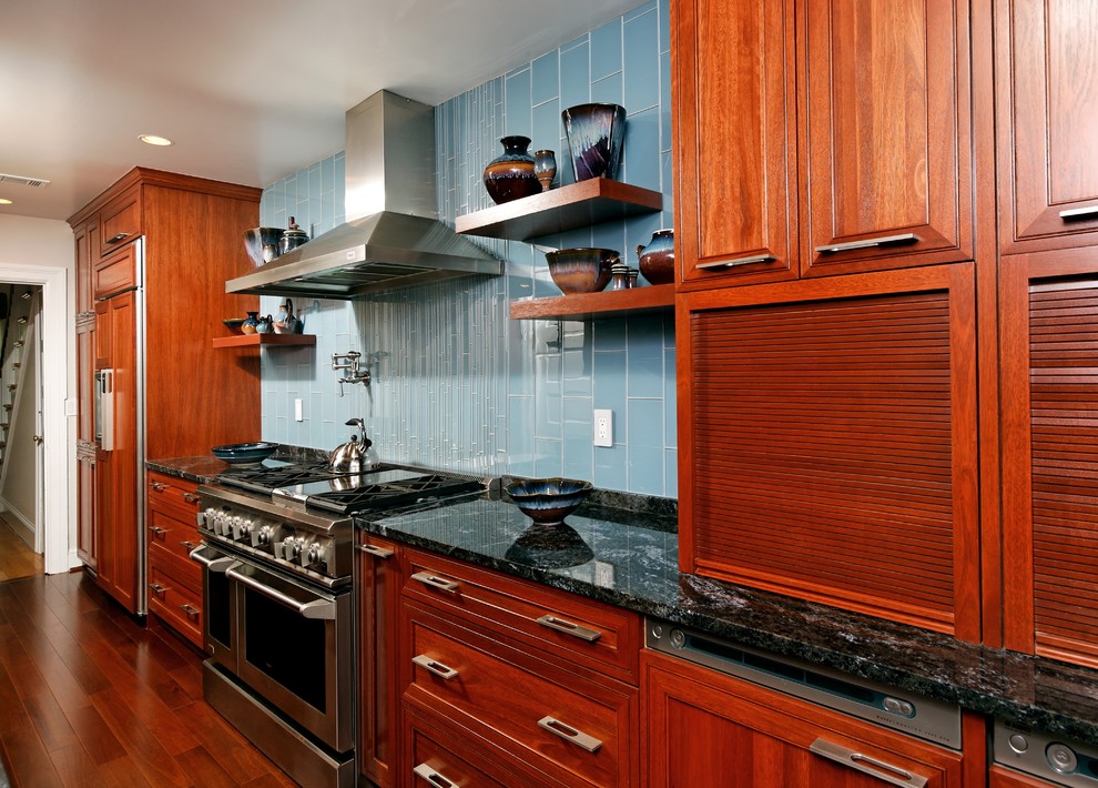 Imagen de cocina actual con encimera de granito, electrodomésticos con paneles, armarios con paneles con relieve, puertas de armario de madera en tonos medios, salpicadero azul y salpicadero de azulejos de vidrio
