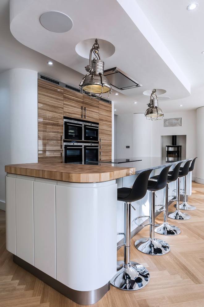 Inspiration for a large contemporary galley open plan kitchen in Essex with a submerged sink, flat-panel cabinets, white cabinets, quartz worktops, black appliances, light hardwood flooring and an island.