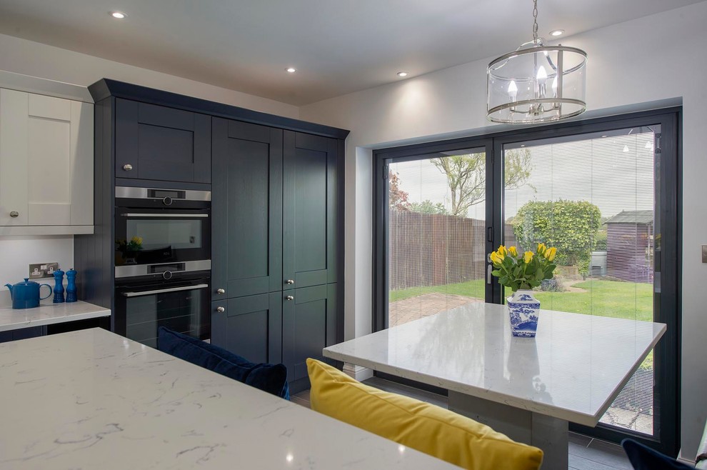 Design ideas for a medium sized contemporary l-shaped kitchen/diner in Other with a submerged sink, shaker cabinets, blue cabinets, quartz worktops, grey splashback, glass sheet splashback, stainless steel appliances, porcelain flooring, a breakfast bar, grey floors and white worktops.