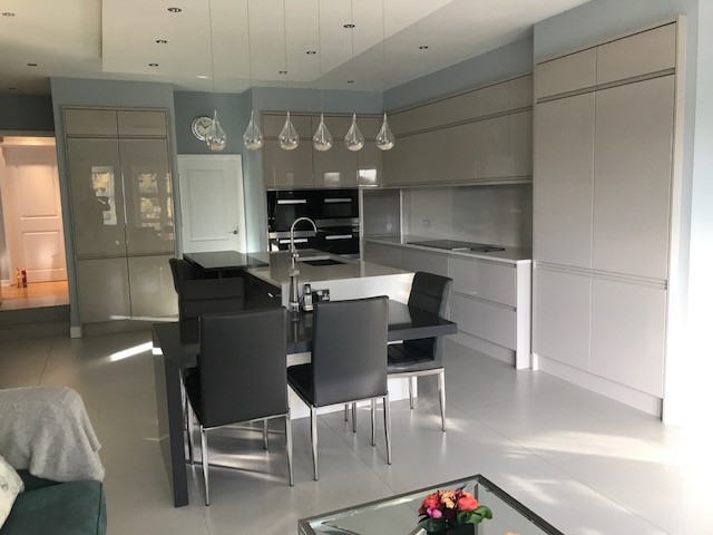 Design ideas for a large modern l-shaped open plan kitchen in Essex with flat-panel cabinets, grey cabinets, quartz worktops, white splashback, glass sheet splashback, stainless steel appliances and an island.