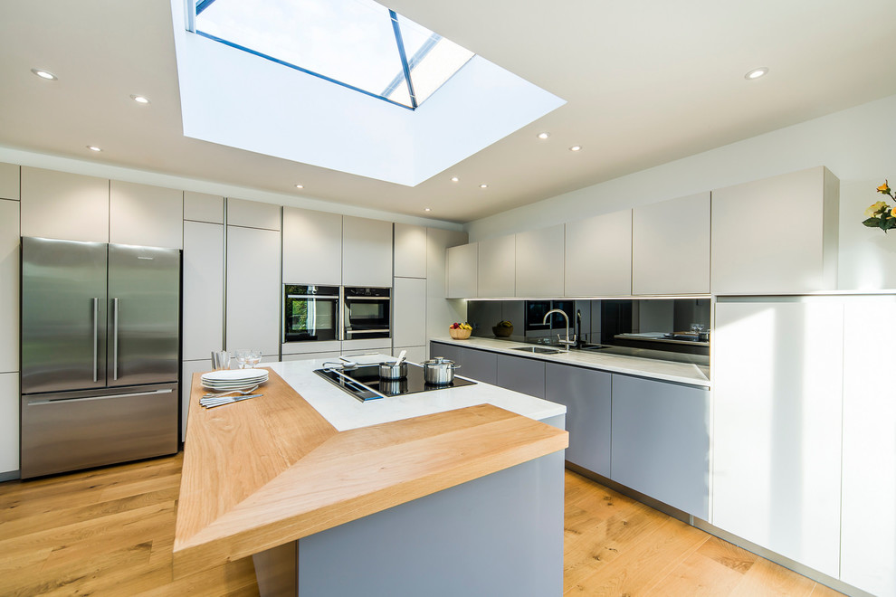 Large contemporary l-shaped open plan kitchen in Wiltshire with flat-panel cabinets, grey cabinets, composite countertops, grey splashback, mirror splashback, black appliances, medium hardwood flooring and an island.