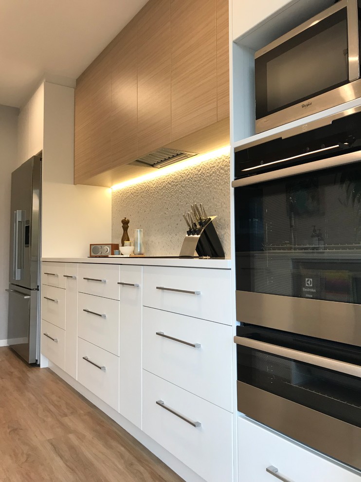 Large scandi galley enclosed kitchen in Christchurch with a double-bowl sink, flat-panel cabinets, white cabinets, engineered stone countertops, grey splashback, porcelain splashback, stainless steel appliances, an island, brown floors and vinyl flooring.