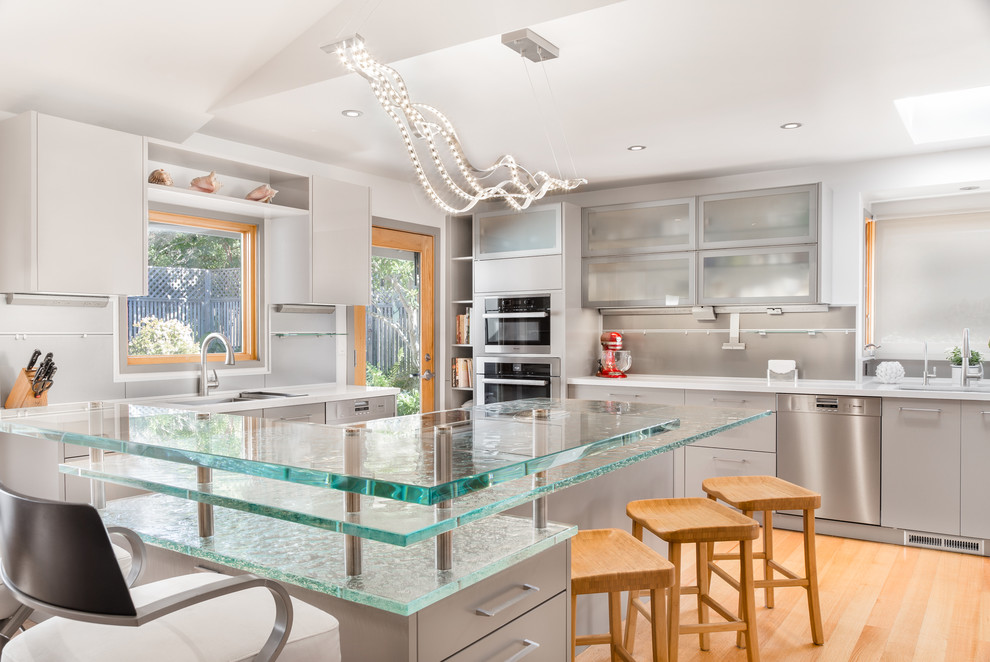 Bild på ett stort funkis kök, med släta luckor, beige skåp, bänkskiva i glas, stänkskydd med metallisk yta, rostfria vitvaror, ljust trägolv och flera köksöar