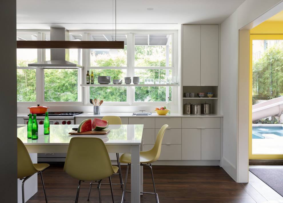 На фото: параллельная кухня у окна в современном стиле с плоскими фасадами, белыми фасадами, техникой из нержавеющей стали, темным паркетным полом, белой столешницей и обеденным столом