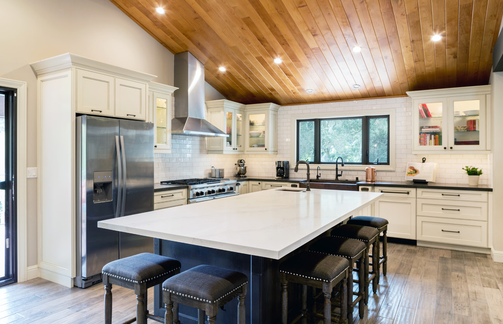 Large rural l-shaped open plan kitchen in San Francisco with a belfast sink, recessed-panel cabinets, white cabinets, engineered stone countertops, white splashback, metro tiled splashback, stainless steel appliances, laminate floors, an island and beige floors.