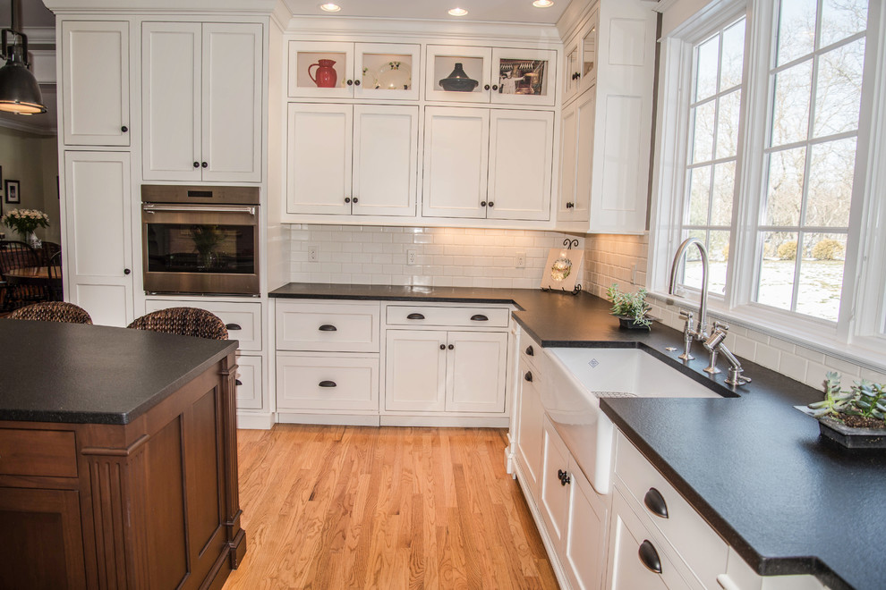 Photo of a large classic u-shaped kitchen/diner in Grand Rapids with a belfast sink, recessed-panel cabinets, white cabinets, granite worktops, white splashback, metro tiled splashback, stainless steel appliances, light hardwood flooring and an island.