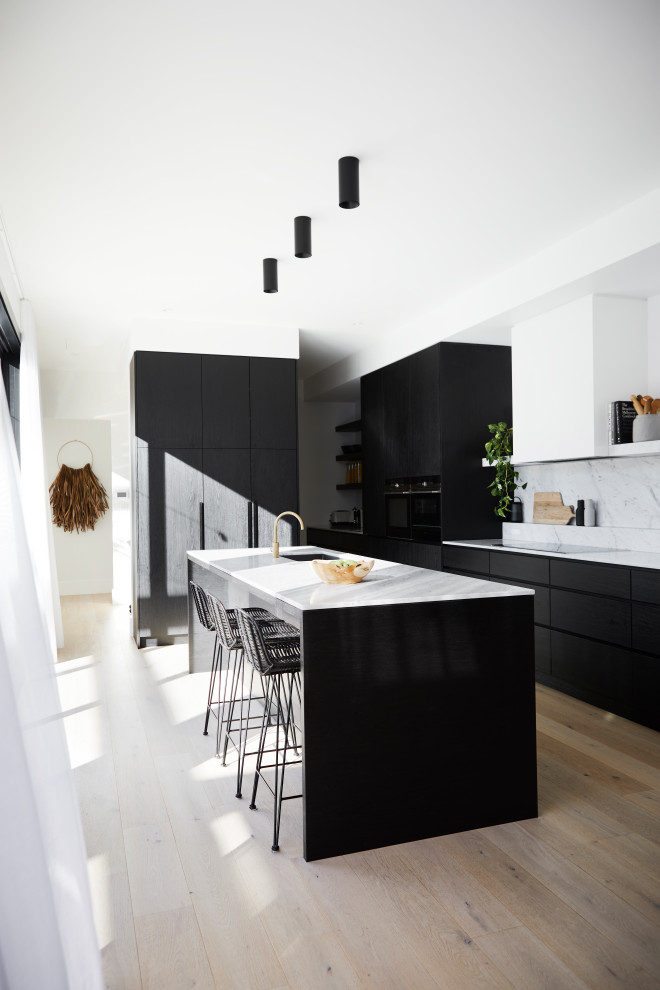 Foto på ett mellanstort funkis vit linjärt kök med öppen planlösning, med en undermonterad diskho, släta luckor, svarta skåp, marmorbänkskiva, vitt stänkskydd, stänkskydd i marmor, svarta vitvaror, ljust trägolv, en köksö och beiget golv