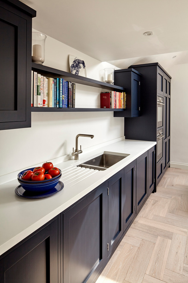 Medium sized contemporary single-wall open plan kitchen in London with a submerged sink, shaker cabinets, blue cabinets, composite countertops, light hardwood flooring, an island and white worktops.