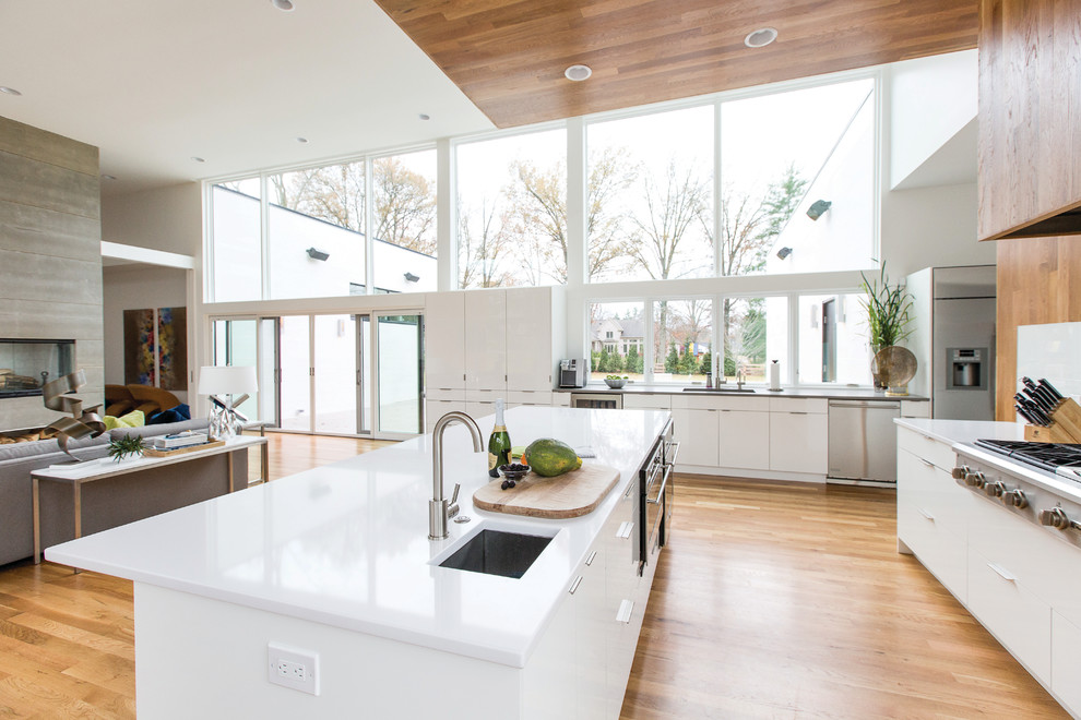Inspiration for a large contemporary l-shaped open plan kitchen in Cincinnati with a submerged sink, flat-panel cabinets, white cabinets, composite countertops, white splashback, glass sheet splashback, stainless steel appliances, medium hardwood flooring, an island, brown floors and white worktops.