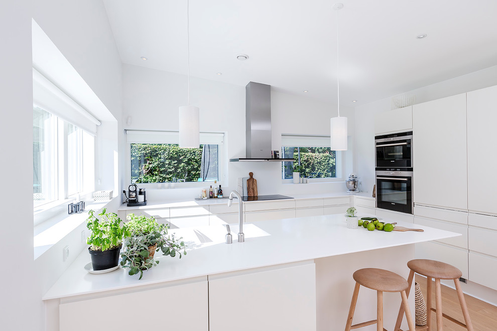 Foto på ett mellanstort funkis kök, med släta luckor, vita skåp, vitt stänkskydd, rostfria vitvaror, ljust trägolv, en halv köksö, en integrerad diskho och laminatbänkskiva
