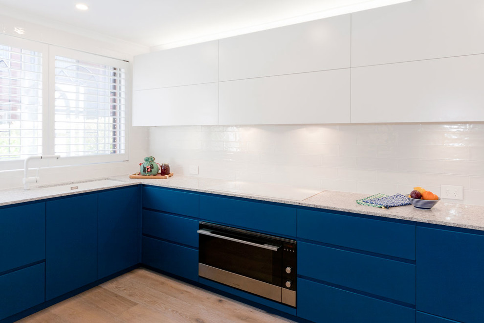Medium sized contemporary l-shaped kitchen in Sydney with flat-panel cabinets, blue cabinets, limestone worktops, white splashback, metro tiled splashback, stainless steel appliances and light hardwood flooring.