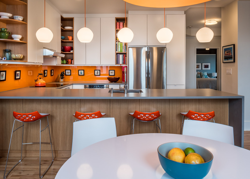 Foto på ett funkis u-kök, med en undermonterad diskho, släta luckor, vita skåp, orange stänkskydd, rostfria vitvaror, ljust trägolv och en halv köksö