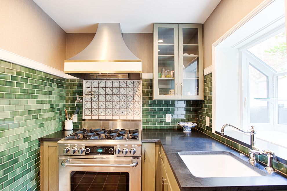 Klassisk inredning av ett litet, avskilt l-kök, med en undermonterad diskho, luckor med glaspanel, grönt stänkskydd, stänkskydd i tunnelbanekakel, rostfria vitvaror, gröna skåp, granitbänkskiva och klinkergolv i porslin
