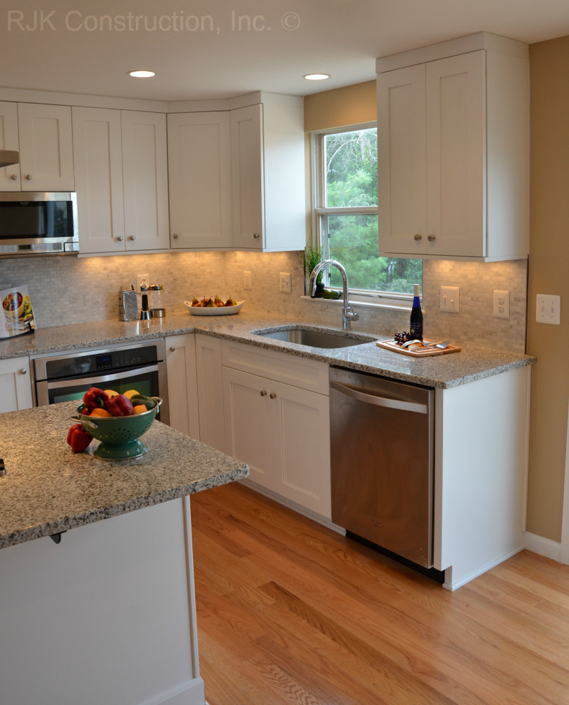 Expansive classic l-shaped open plan kitchen in DC Metro with a submerged sink, shaker cabinets, white cabinets, granite worktops, grey splashback, ceramic splashback, stainless steel appliances, light hardwood flooring and an island.