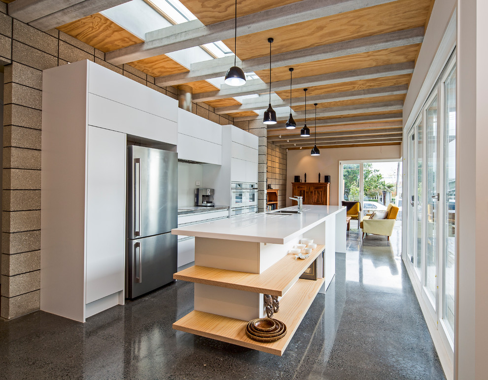 This is an example of a medium sized industrial galley open plan kitchen in Auckland with a submerged sink, flat-panel cabinets, white cabinets, beige splashback, glass sheet splashback, stainless steel appliances, concrete flooring, an island, grey floors and white worktops.