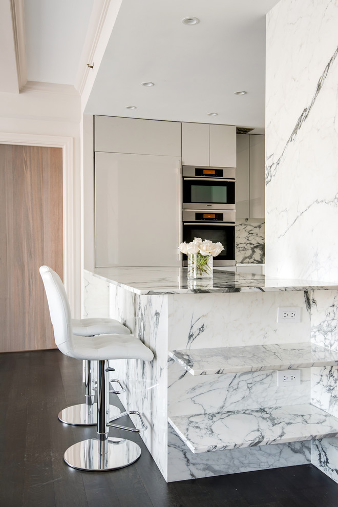 Inspiration for a medium sized contemporary u-shaped kitchen in New York with a submerged sink, flat-panel cabinets, white cabinets, marble worktops, white splashback, stone slab splashback, integrated appliances, dark hardwood flooring, a breakfast bar, brown floors and white worktops.