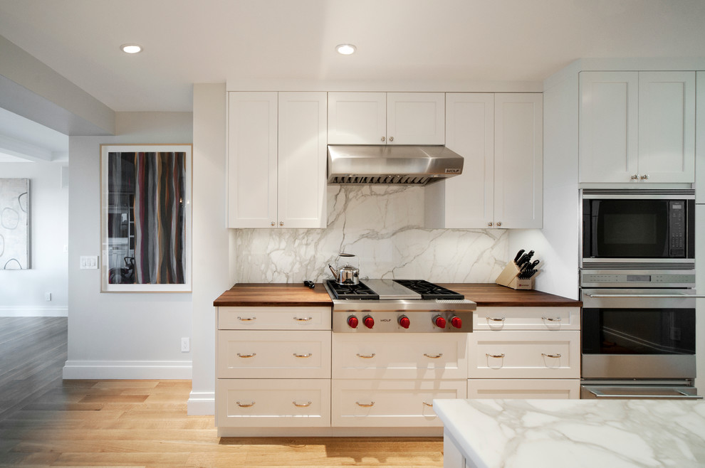 Example of a mid-sized trendy light wood floor and beige floor open concept kitchen design in New York with an undermount sink, glass-front cabinets, white cabinets, marble countertops, white backsplash, marble backsplash, stainless steel appliances and a peninsula