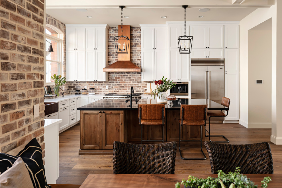 Photo of a large traditional l-shaped kitchen/diner in Phoenix with a submerged sink, shaker cabinets, white cabinets, engineered stone countertops, brown splashback, brick splashback, stainless steel appliances, light hardwood flooring, an island, beige floors and black worktops.