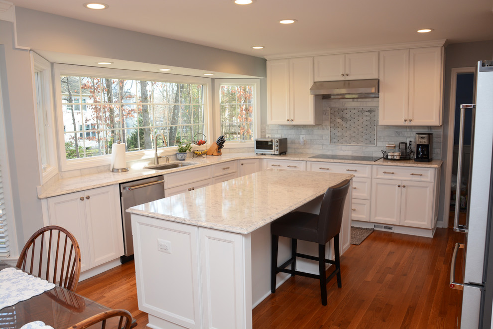 Klassisk inredning av ett mellanstort vit vitt kök, med en enkel diskho, luckor med infälld panel, vita skåp, bänkskiva i kvarts, grått stänkskydd, rostfria vitvaror, mellanmörkt trägolv, en köksö och brunt golv