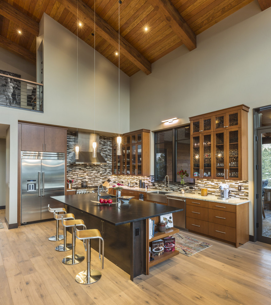 Modelo de cocina actual con fregadero sobremueble, armarios tipo vitrina, puertas de armario de madera oscura, salpicadero multicolor, salpicadero de azulejos en listel, electrodomésticos de acero inoxidable, suelo de madera en tonos medios, una isla, suelo marrón y encimeras beige