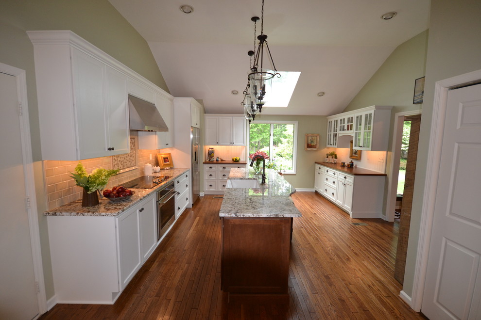Example of a large transitional l-shaped medium tone wood floor enclosed kitchen design in Philadelphia with a farmhouse sink, recessed-panel cabinets, white cabinets, granite countertops, beige backsplash, subway tile backsplash, stainless steel appliances and an island