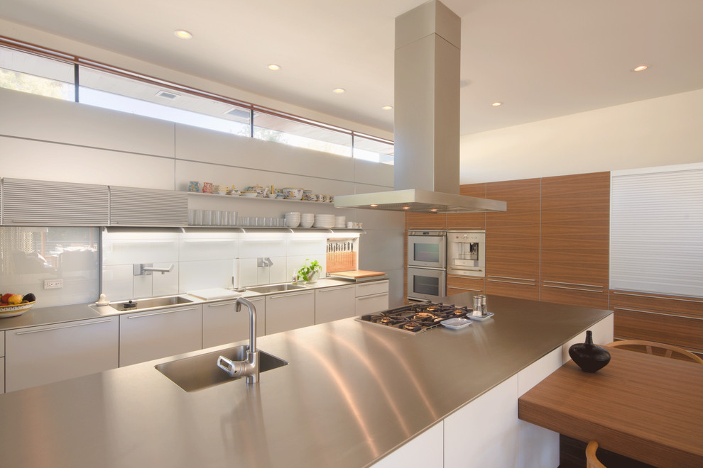 Bild på ett stort funkis kök, med en integrerad diskho, bänkskiva i rostfritt stål, släta luckor, vita skåp, rostfria vitvaror, vitt stänkskydd, glaspanel som stänkskydd, en köksö och ljust trägolv