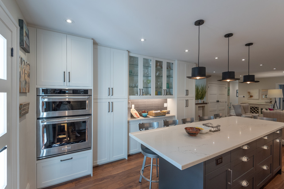 Inspiration for a medium sized farmhouse single-wall open plan kitchen in Toronto with a double-bowl sink, shaker cabinets, white cabinets, marble worktops, white splashback, metro tiled splashback, stainless steel appliances, dark hardwood flooring, an island, brown floors and white worktops.