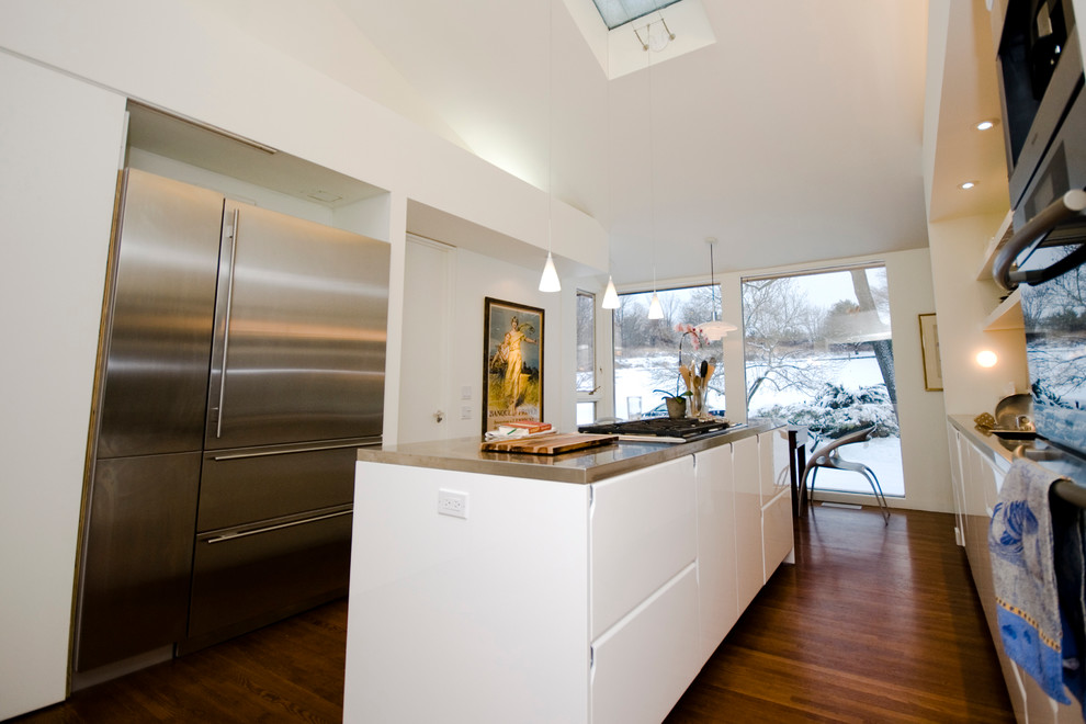 Foto på ett stort funkis linjärt kök och matrum, med en undermonterad diskho, släta luckor, bänkskiva i rostfritt stål, grått stänkskydd, rostfria vitvaror, mellanmörkt trägolv, en köksö, vita skåp och stänkskydd i metallkakel