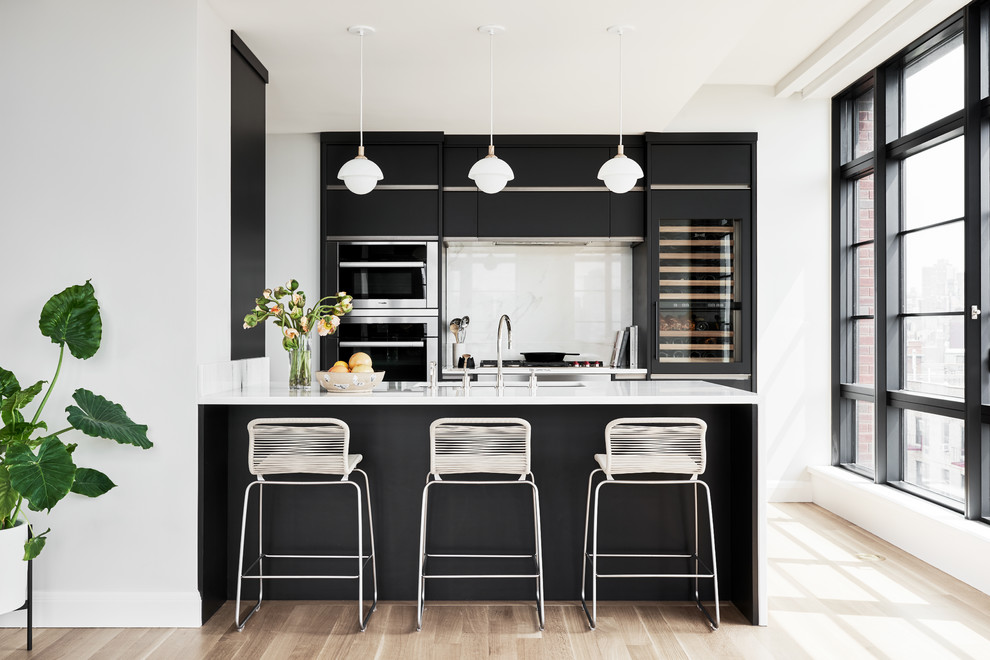 Bild på ett minimalistiskt vit vitt kök, med en undermonterad diskho, släta luckor, svarta skåp, vitt stänkskydd, glaspanel som stänkskydd, rostfria vitvaror, ljust trägolv och en halv köksö