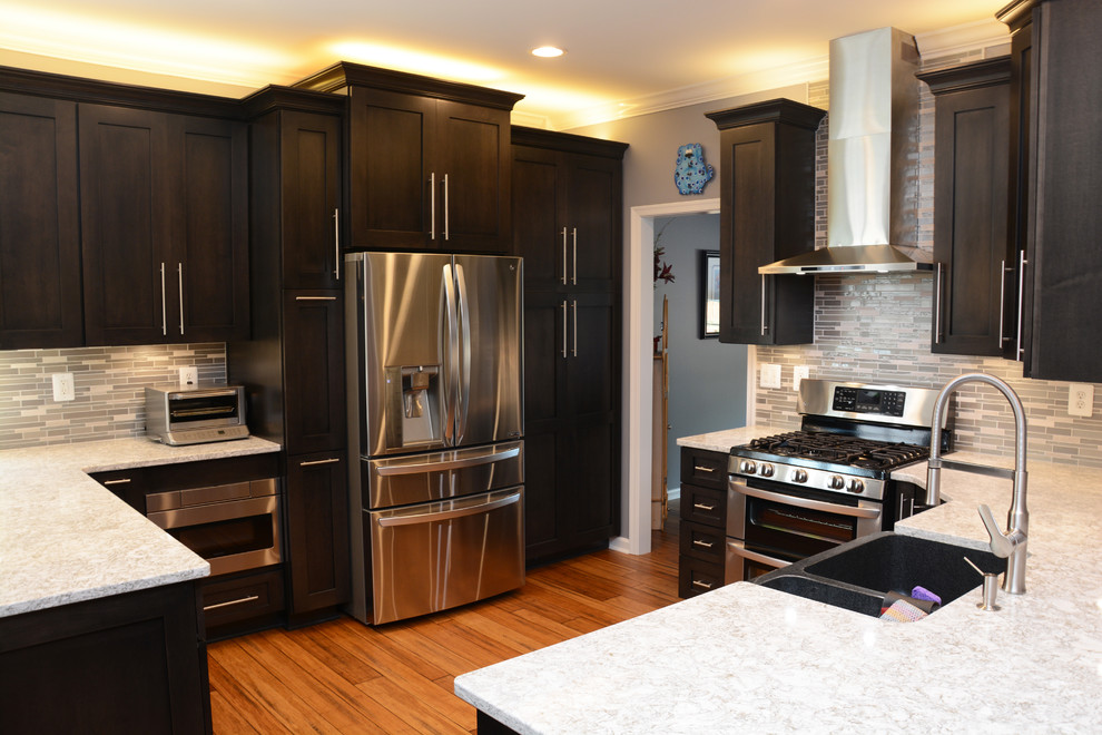 This is an example of a medium sized classic u-shaped open plan kitchen in DC Metro with a belfast sink, recessed-panel cabinets, black cabinets, engineered stone countertops, grey splashback, mosaic tiled splashback, stainless steel appliances, light hardwood flooring, a breakfast bar and brown floors.