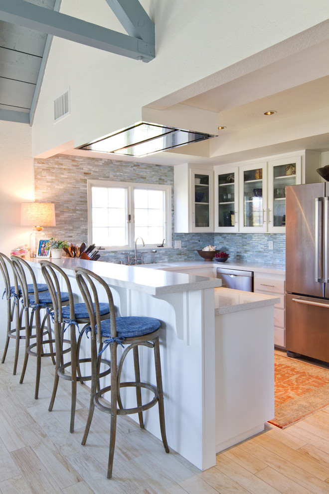 Beach style u-shaped light wood floor kitchen photo in Los Angeles with an undermount sink, glass-front cabinets, white cabinets, blue backsplash, mosaic tile backsplash and stainless steel appliances