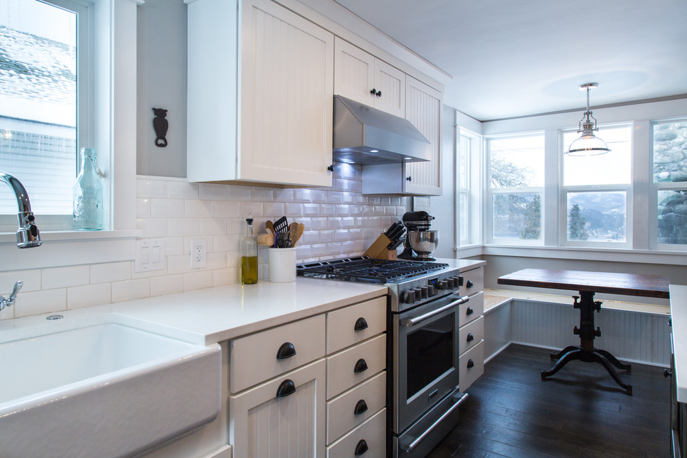 Small country galley kitchen/diner in Portland with a belfast sink, shaker cabinets, white cabinets, engineered stone countertops, white splashback, metro tiled splashback, stainless steel appliances, dark hardwood flooring and an island.