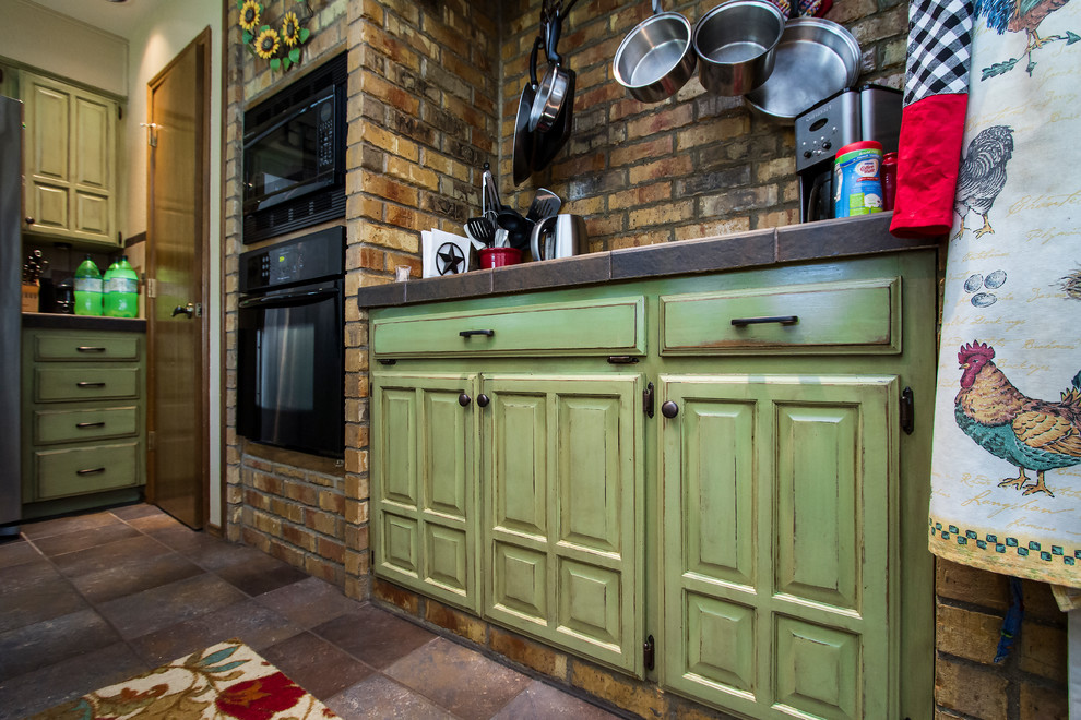 Inspiration for a medium sized shabby-chic style l-shaped kitchen/diner in Austin with raised-panel cabinets, green cabinets, tile countertops, brown splashback, black appliances, ceramic flooring and brick splashback.