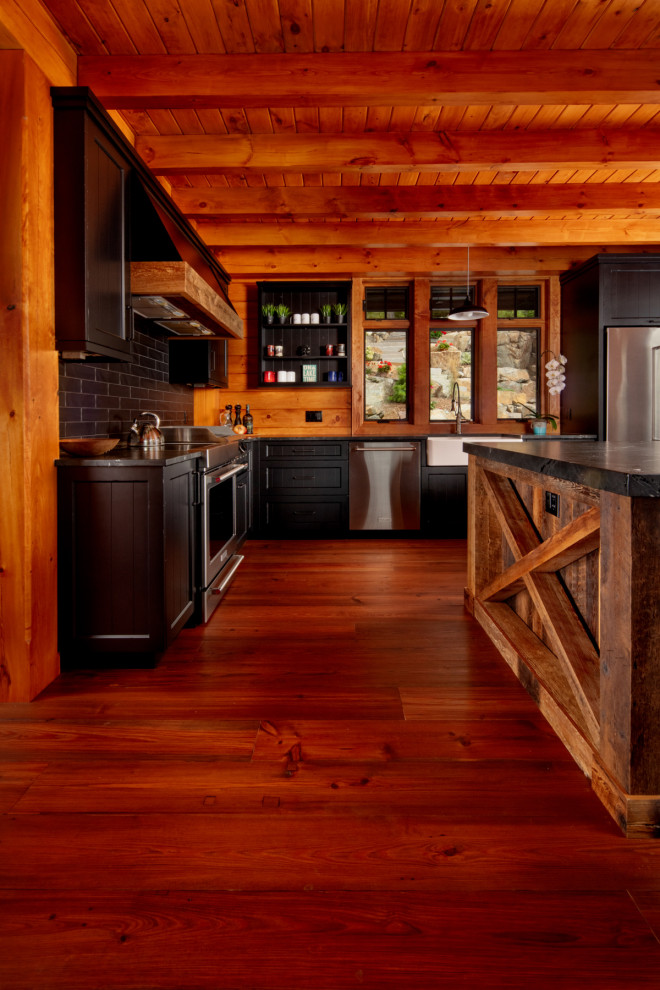 Example of a mid-sized mountain style l-shaped medium tone wood floor and brown floor eat-in kitchen design in Other with a farmhouse sink, shaker cabinets, black cabinets, quartz countertops, black backsplash, stainless steel appliances, an island and black countertops