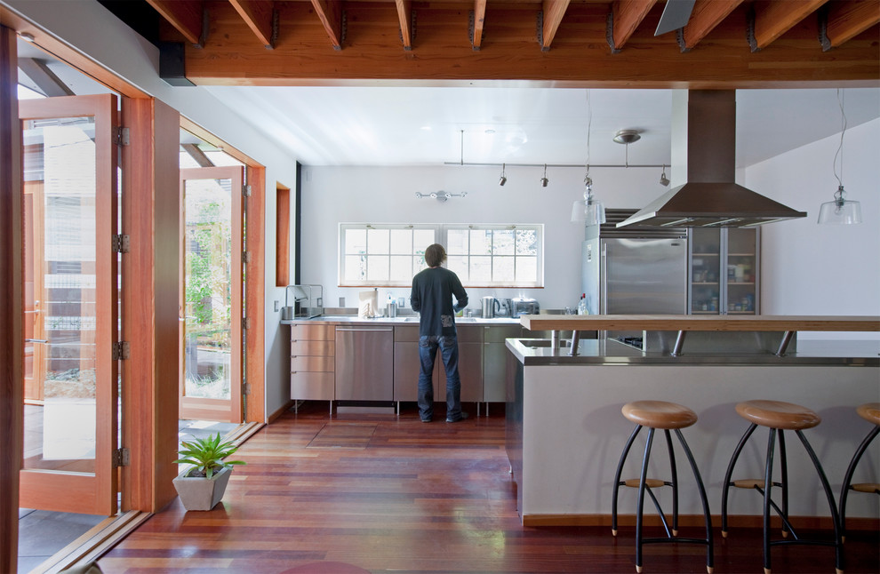 Modern inredning av ett mellanstort linjärt kök med öppen planlösning, med luckor med glaspanel, skåp i rostfritt stål, bänkskiva i rostfritt stål, rostfria vitvaror, mellanmörkt trägolv, en undermonterad diskho, en halv köksö och brunt golv