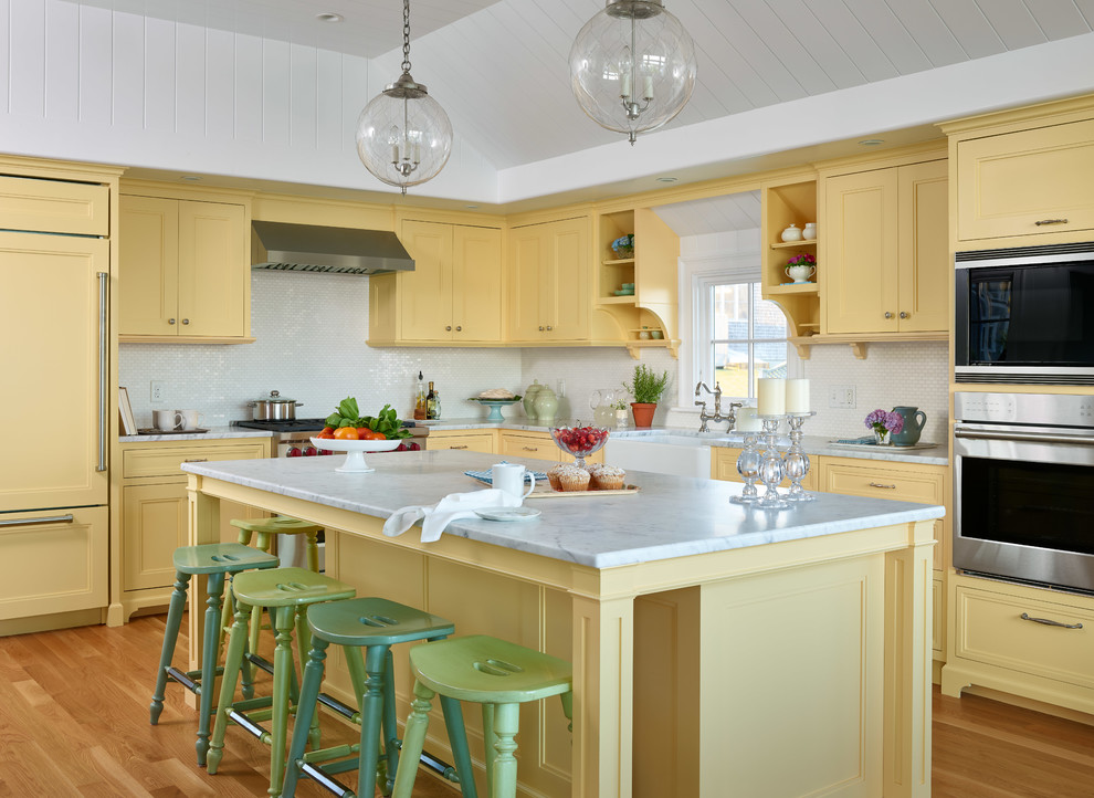 Kitchen - coastal l-shaped light wood floor kitchen idea in Boston with a farmhouse sink, yellow cabinets, marble countertops, white backsplash, paneled appliances, an island, white countertops and recessed-panel cabinets