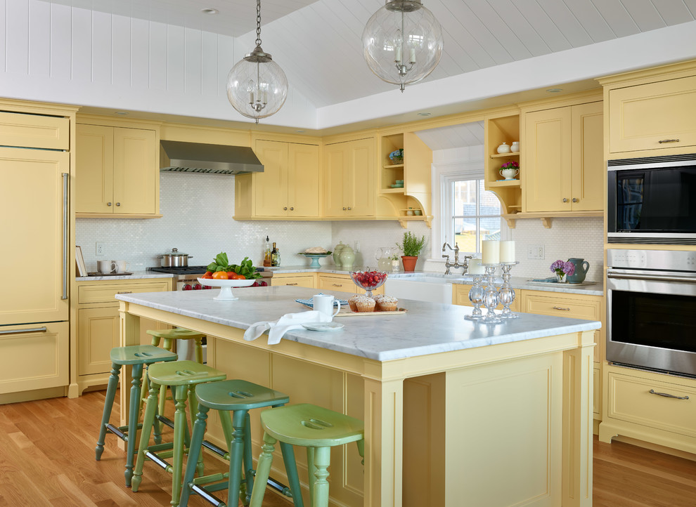 Coastal l-shaped kitchen in Boston with a belfast sink, yellow cabinets, marble worktops, white splashback, integrated appliances, light hardwood flooring, an island, white worktops and recessed-panel cabinets.
