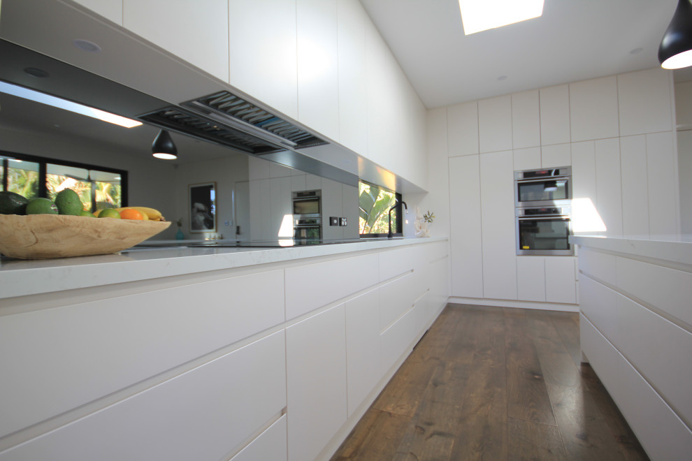 Bild på ett mellanstort funkis vit vitt kök, med en undermonterad diskho, vita skåp, bänkskiva i kvarts, glaspanel som stänkskydd, rostfria vitvaror, en köksö och brunt golv