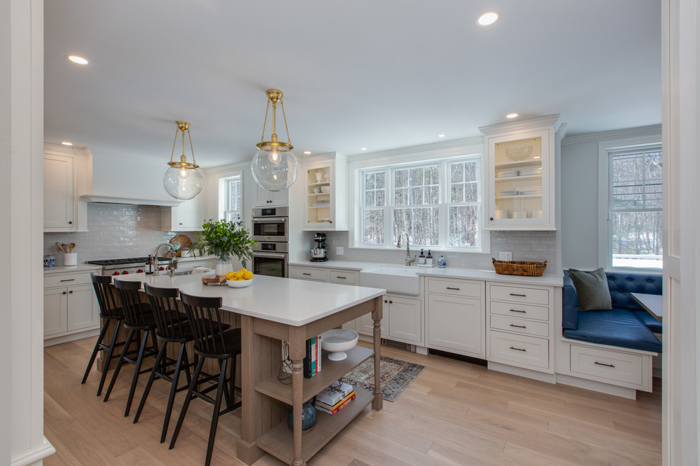 Inspiration for a mid-sized transitional l-shaped medium tone wood floor and brown floor eat-in kitchen remodel in Boston with a farmhouse sink, beaded inset cabinets, white cabinets, quartz countertops, gray backsplash, subway tile backsplash, stainless steel appliances, an island and white countertops