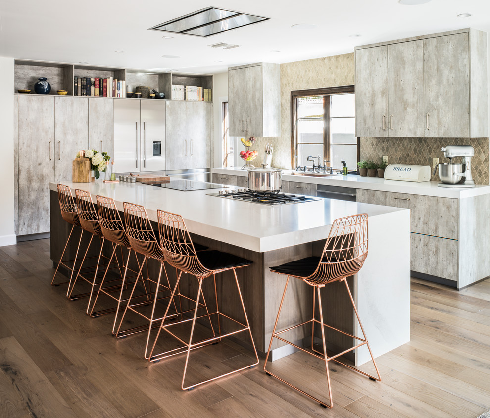 Inspiration for a large contemporary l-shaped open plan kitchen in Los Angeles with a submerged sink, flat-panel cabinets, beige splashback, stone tiled splashback, stainless steel appliances, light hardwood flooring, an island, distressed cabinets and composite countertops.