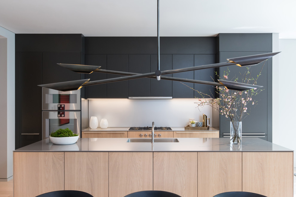 This is an example of a medium sized contemporary galley kitchen/diner in New York with flat-panel cabinets, an island, a submerged sink, black cabinets, white splashback, light hardwood flooring, beige floors and grey worktops.