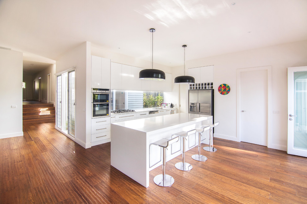 Große, Offene Moderne Küche in L-Form mit Doppelwaschbecken, weißen Schränken, Glasrückwand, Küchengeräten aus Edelstahl, braunem Holzboden, Kücheninsel und flächenbündigen Schrankfronten in Melbourne