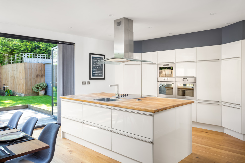 Photo of a large contemporary single-wall open plan kitchen in Gloucestershire with flat-panel cabinets, white cabinets, stainless steel appliances, light hardwood flooring, an island, a built-in sink and wood worktops.