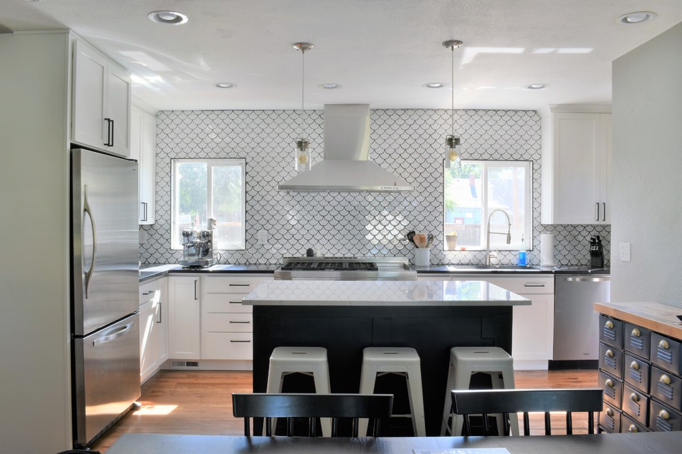 This is an example of a medium sized farmhouse l-shaped kitchen/diner in Denver with a submerged sink, shaker cabinets, white cabinets, engineered stone countertops, white splashback, mosaic tiled splashback, stainless steel appliances, medium hardwood flooring, an island, brown floors and black worktops.