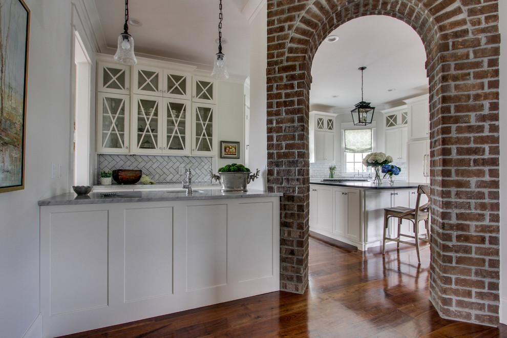 Example of a large classic medium tone wood floor kitchen design in Nashville with a farmhouse sink, shaker cabinets, white cabinets, marble countertops, gray backsplash, stone tile backsplash, paneled appliances and an island
