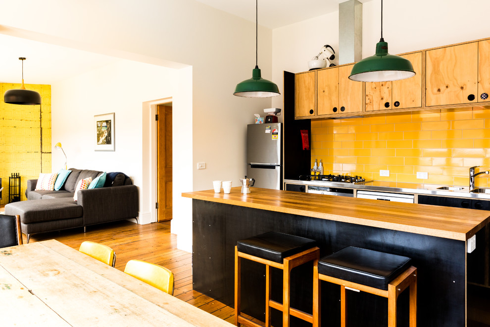Modern inredning av ett kök, med en integrerad diskho, skåp i ljust trä, bänkskiva i rostfritt stål, gult stänkskydd, stänkskydd i keramik, rostfria vitvaror, mellanmörkt trägolv, en köksö och brunt golv