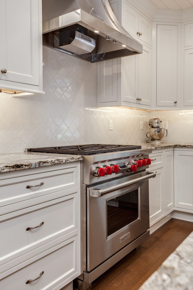 Cette photo montre une cuisine américaine chic en U de taille moyenne avec un évier encastré, un placard à porte shaker, des portes de placard blanches, un plan de travail en granite, une crédence blanche, une crédence en céramique, un électroménager en acier inoxydable, un sol en bois brun, îlot, un sol marron et un plan de travail gris.