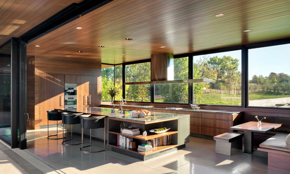 На фото: кухня у окна в современном стиле с обеденным столом, плоскими фасадами, фасадами цвета дерева среднего тона, техникой под мебельный фасад, бетонным полом, островом, серым полом и серой столешницей