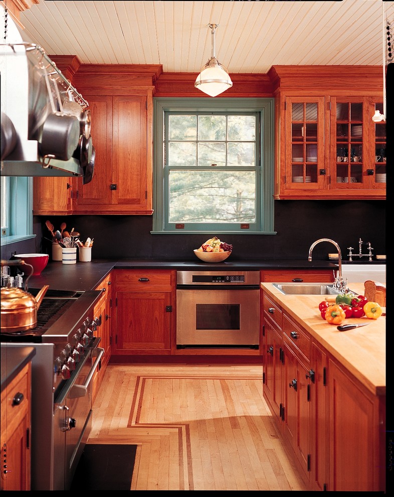 Inredning av ett amerikanskt avskilt, mellanstort l-kök, med en rustik diskho, skåp i shakerstil, skåp i mellenmörkt trä, granitbänkskiva, svart stänkskydd, stänkskydd i sten, rostfria vitvaror och ljust trägolv